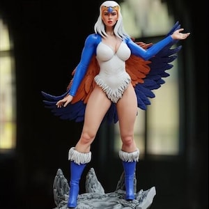 以下が含まれることがあります： 羽根の冠と羽根のついた翼を持つ白と青のフィギュアが、岩の台座の上に立っています。フィギュアは、青いマントと茶色の羽根の翼が付いた白いボディスーツを着ています。台座には「Masters of the Universe」の文字が書かれています。