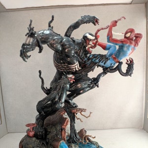 May include: A black and red action figure of Venom battling Spider-Man. The figures are posed in a dynamic fight scene on a base with a blue water effect and a brown tree trunk.