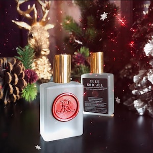May include: Two frosted glass perfume bottles with gold caps. One bottle has a red wax seal with the letter "R". The other bottle has a label that reads "Yule God Jul". The background includes Christmas decorations.