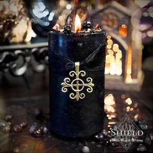 May include: A cylindrical black pillar candle with a lit flame, topped with small dark stones. A black ribbon is tied around the center, holding a gold-toned metal cross pendant with ornate, swirling filigree details. The word SHIELD is visible.
