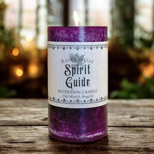 May include: A lit, purple intention candle with a label that reads "Raven Spells Spirit Guide Intention Candle Old World Magick." The candle is cylindrical and sits on a wooden surface. The flame is bright.