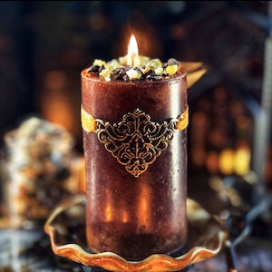 May include: A lit brown pillar candle with a decorative gold filigree band and a yellow ribbon. The top of the candle is adorned with colorful crystals. The candle sits on a decorative gold-colored dish.