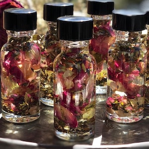 May include: Several clear glass bottles filled with a liquid and botanical elements, including pink rose petals and green herbs. Each bottle has a black cap. The bottles are arranged on a silver tray.