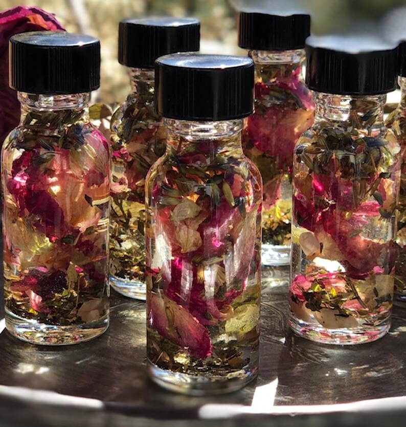 May include: Several clear glass bottles filled with a liquid and botanical elements, including pink rose petals and green herbs. Each bottle has a black cap. The bottles are arranged on a silver tray.