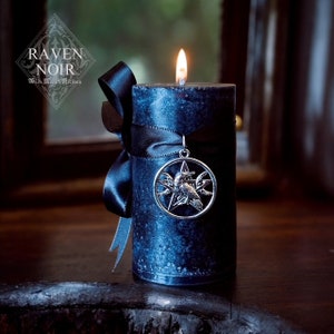 May include: A dark blue candle with a black ribbon and a silver charm featuring a raven and a pentagram. The candle is lit and the flame is visible. The text "Raven Noir" is visible on the label.