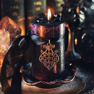 May include: A lit black pillar candle with a purple top, adorned with a black ribbon and a gold-colored filigree pendant. The candle sits on a decorative dish, creating a gothic or mystical aesthetic.