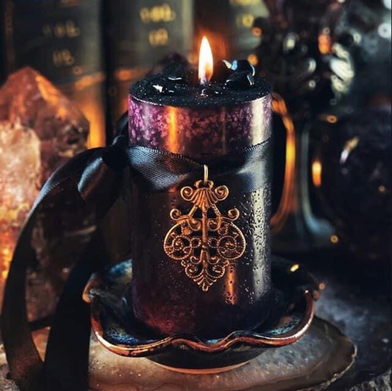 May include: A lit black pillar candle with a purple top, adorned with a black ribbon and a gold-colored filigree pendant. The candle sits on a decorative dish, creating a gothic or mystical aesthetic.