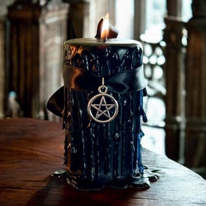 May include: A black pillar candle with a ribbon and a pentagram charm. The candle has a textured, dripped wax appearance and is lit with a flame. The candle is on a wooden surface.