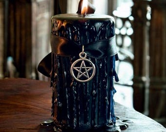 May include: A black pillar candle with a ribbon and a pentagram charm. The candle has a textured, dripped wax appearance and is lit with a flame. The candle is on a wooden surface.