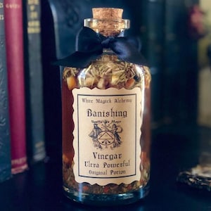 May include: A glass bottle with a cork stopper and a black ribbon tied around the neck. The bottle is filled with a brown liquid and herbs. The label reads "White Magick Alchemy Banishing Spells for Magic Vinegar Ultra Powerful Original Potion".