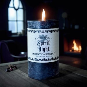 May include: A burning dark blue pillar candle with a white label. The label reads "Raven Spells Spirit Night Intention Candle Old World Magick." The candle sits on a wooden surface, with a blurred window and fireplace in the background.
