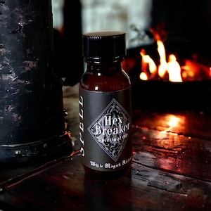 May include: A small brown glass bottle with a black cap and a black and white label that reads "Hex Breaker Spiritual Oil" and "White Magick Alchemy". The bottle is sitting on a wooden surface in front of a fireplace.