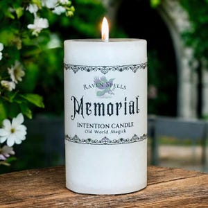 May include: A white, cylindrical candle with a lit wick. The candle has a label with the words "Raven Spells Memorial Intention Candle Old World Magick" in black script and decorative borders. The candle sits on a wooden surface.