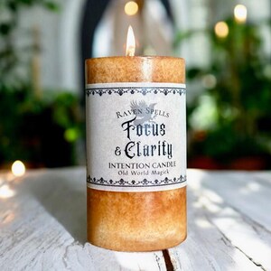 May include: A lit, cylindrical intention candle with a label that reads "Raven Spells Focus & Clarity." The candle has a textured, light brown exterior and a white label with black text and decorative borders. The flame is bright and the candle is on a white wooden surface.