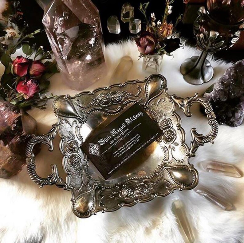 Pu&ograve; includere: Vassoio argentato ornato con dettagli floreali e volute, contenente un biglietto da visita. Il biglietto da visita recita "White Magick Alchemy". Il vassoio &egrave; su una superficie bianca simile a pelliccia, circondato da cristalli e fiori secchi.
