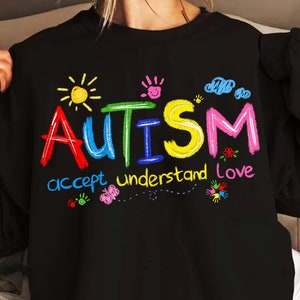 May include: Black sweatshirt with the word "AUTISM" written in colorful letters, with the words "accept understand love" written below. The letters are drawn in a childlike style with colorful drawings of a sun, clouds, and butterflies.