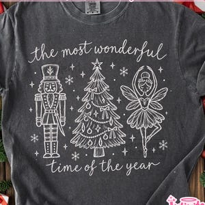 May include: A charcoal grey t-shirt featuring a white line art graphic of a nutcracker, a decorated Christmas tree, and a ballerina. The text reads, "the most wonderful time of the year." The shirt has a crew neckline and short sleeves.