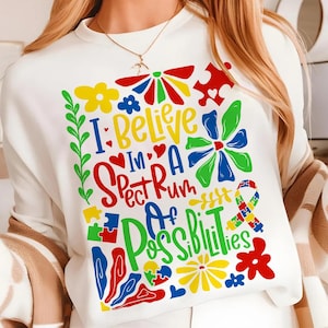 May include: White sweatshirt with a colorful graphic design that says "I believe in a spectrum of possibilities". The design includes flowers, hearts, puzzle pieces, and a rainbow ribbon.