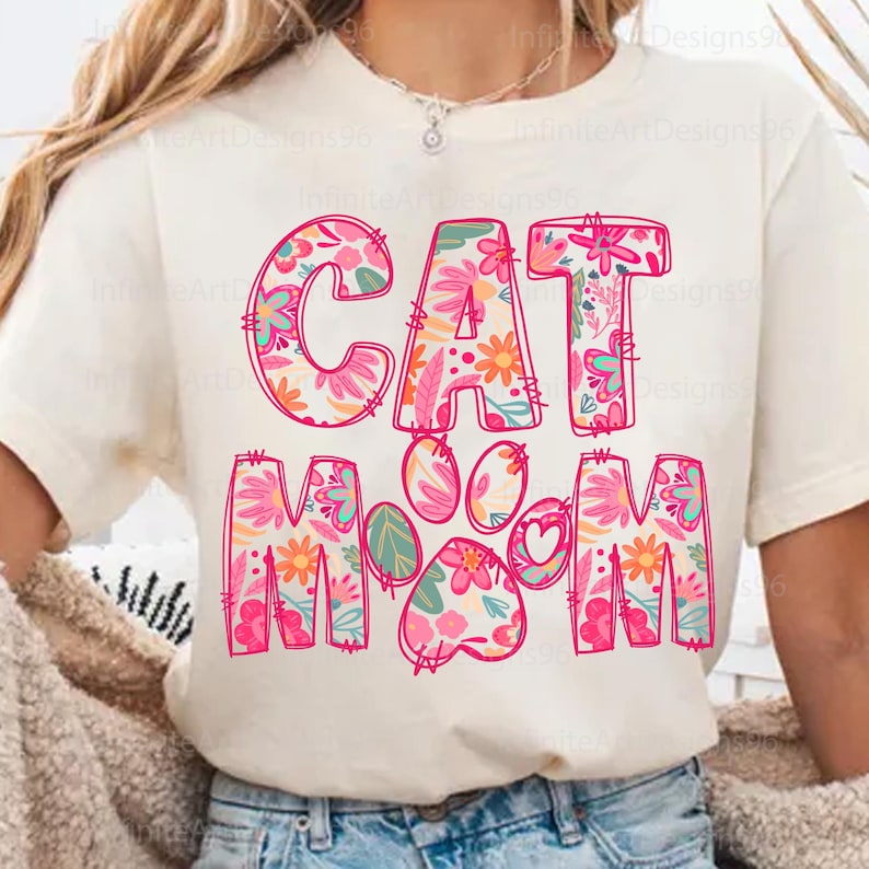 May include: White t-shirt with a pink floral design that says "Cat Mom" with a paw print in the middle.