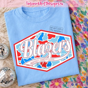 Op de afbeelding: Een lichtblauw t-shirt met een zeshoekige afbeelding in een rood, wit en lichtblauw camouflagepatroon. Het woord "Blazers" staat in grote witte schrijfletters met een rode omlijning boven de tekst "UNIVERSITY CHARTER SCHOOL".
