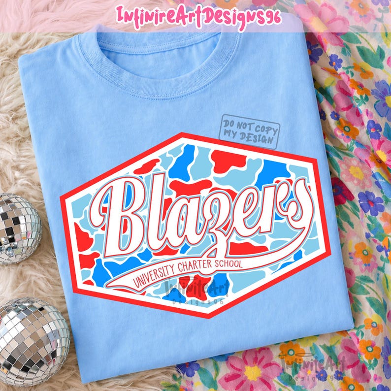 Op de afbeelding: Een lichtblauw t-shirt met een zeshoekige afbeelding in een rood, wit en lichtblauw camouflagepatroon. Het woord "Blazers" staat in grote witte schrijfletters met een rode omlijning boven de tekst "UNIVERSITY CHARTER SCHOOL".