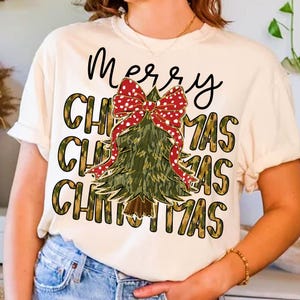 Può includere: T-shirt color crema con un design a forma di albero di Natale. L'albero è verde con un fiocco rosso a pois. Le parole "Merry Christmas" sono scritte in nero sopra l'albero, con "Christmas" ripetuto tre volte in oro.