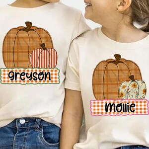 Può includere: Due magliette color crema con grafiche di zucche. Una maglietta presenta una grande zucca a quadri con due zucche più piccole e il nome "greyson" su uno striscione a quadri. L'altra maglietta ha una zucca a quadri e una zucca floreale con il nome "mollie".