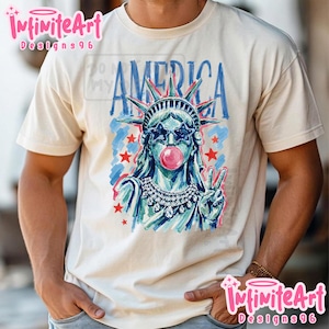 May include: A cream-coloured t-shirt featuring a graphic of the Statue of Liberty wearing star-shaped sunglasses and blowing a pink bubblegum bubble. The word AMERICA is printed in blue above the figure, accompanied by red stars and blue brushstrokes.