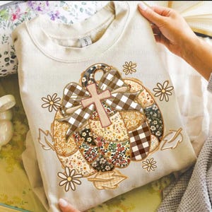 Könnte beinhalten: Cremefarbenes Sweatshirt mit einem Kürbisdesign und einem Kreuz. Der Kürbis besteht aus Patchwork-Stoff in verschiedenen Mustern, darunter Blumenmuster und Karos. Braun- und Beigetöne dominieren das Design, mit dekorativen Blumen und Blättern.