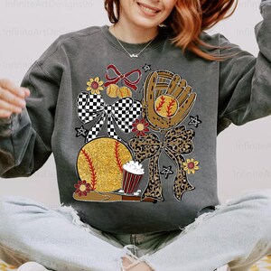 Puede incluir: Sudadera gris oscuro con un diseño temático de béisbol. El diseño incluye una pelota de béisbol dorada con purpurina, un guante de béisbol, lazos, flores y una taza de palomitas de maíz. El texto "Infinite Art Designs96" es visible.