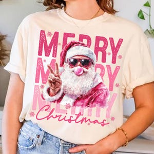 May include: A white t-shirt with a pink graphic of Santa Claus wearing sunglasses and blowing a bubble. The graphic says "Merry Merry Christmas" with snowflakes.