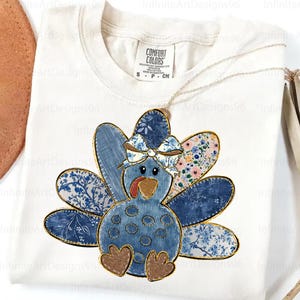 May include: White t-shirt featuring a blue turkey design with floral and denim-like patterns. The turkey has a bow and gold accents. The shirt has a Comfort Colors tag.