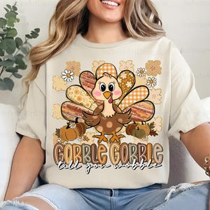 May include: Beige t-shirt featuring a cartoon turkey with patterned feathers, pumpkins, and daisies. The text "GOBBLE GOBBLE till you wobble" is below the turkey. A festive design for Thanksgiving.