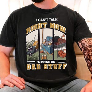 Può includere: T-shirt nera con la scritta "I CAN'T TALK RIGHT NOW I'M DOING HOT DAD STUFF". La grafica raffigura un tosaerba, una scena di pesca e un barbecue.
