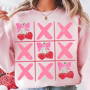 Könnte beinhalten: Hellrosa Sweatshirt mit einem Tic-Tac-Toe-Design. Das Spielbrett hat rosa 'X's und Kirschen mit rosa Schleifen anstelle von 'O's. Das Sweatshirt ist langärmlig und hat einen Rundhalsausschnitt.