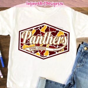 Op de afbeelding: Een wit t-shirt met een zeshoekige afbeelding in een bordeauxrood, goud en wit camouflagepatroon. De tekst "Panthers" is geschreven in een groot, vet, wit scriptlettertype, met "SPRING GARDEN" in een kleiner lettertype op een witte banner.