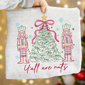 Puede incluir: Sudadera gris claro con un diseño de árbol de Navidad, un lazo y una estrella rosas, flanqueada por dos figuras de cascanueces. La frase "Y'all are nuts" está escrita en rojo. Copos de nieve y estrellas azules dispersos.