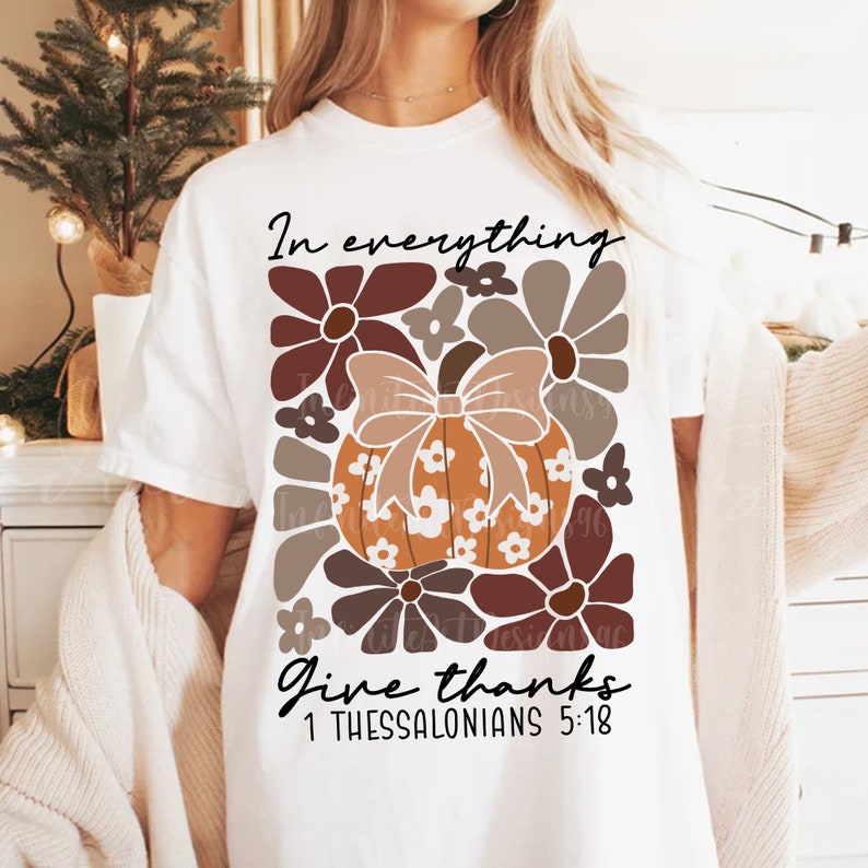 May include: White t-shirt with a graphic design of a pumpkin with a bow and flowers. The text "In everything give thanks 1 Thessalonians 5:18" is printed on the shirt.