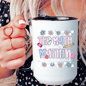 May include: White ceramic mug with a black rim and handle. The mug features the text "The Math Ain't Mathin'" in a colorful, playful font, along with illustrations of school supplies and flowers. The mug is held by a person.