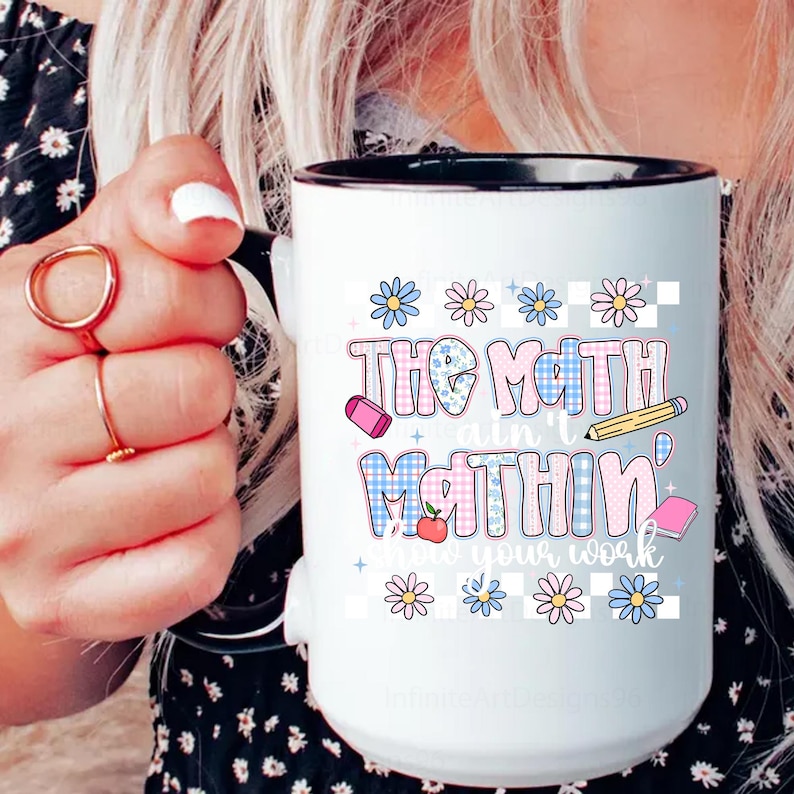 May include: White ceramic mug with a black rim and handle. The mug features the text "The Math Ain't Mathin'" in a colorful, playful font, along with illustrations of school supplies and flowers. The mug is held by a person.