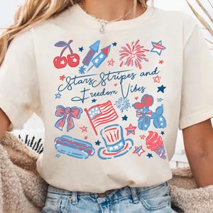 May include: Cream-colored t-shirt with a patriotic design featuring cherries, fireworks, stars, a bow, an American flag, a hot dog, a top hat, an ice cream cone, and the text "Stars Stripes and Freedom Vibes."