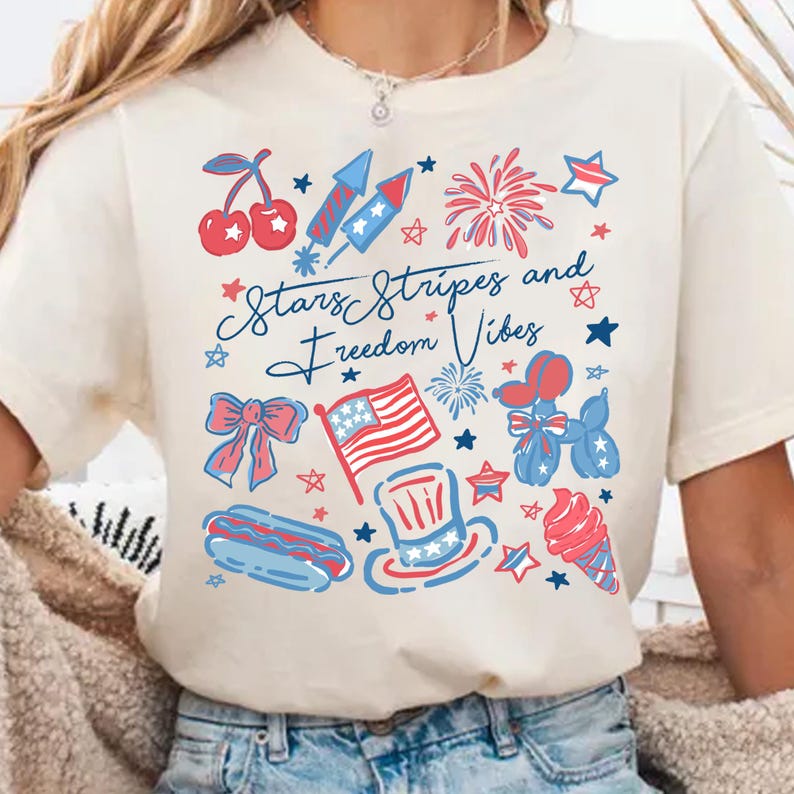May include: Cream-colored t-shirt with a patriotic design featuring cherries, fireworks, stars, a bow, an American flag, a hot dog, a top hat, an ice cream cone, and the text "Stars Stripes and Freedom Vibes."