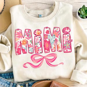May include: A white sweatshirt with the word "MIMI" in pink letters with a floral pattern. The letters are outlined in pink and have a pink bow underneath.