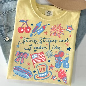 May include: A yellow t-shirt with a patriotic design featuring cherries, fireworks, an American flag, and the text "Stars Stripes and Freedom Vibes." The design includes red, white, and blue elements.