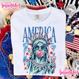 May include: A white t-shirt featuring a graphic of the Statue of Liberty wearing star-shaped sunglasses and blowing a pink bubble. The word AMERICA is printed above in blue. The statue wears a crystal necklace and makes a peace sign gesture.