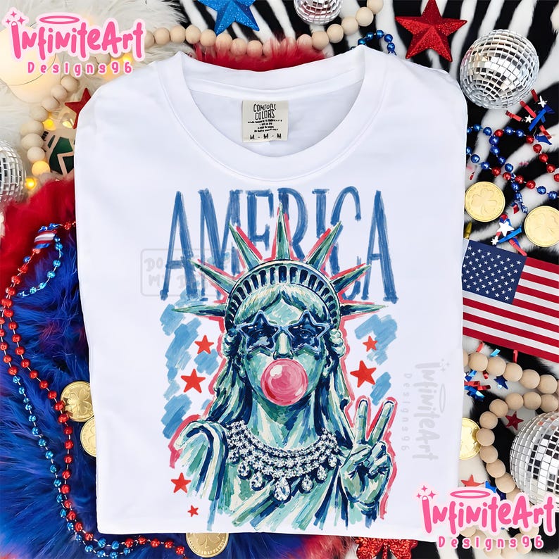 May include: A white t-shirt featuring a graphic of the Statue of Liberty wearing star-shaped sunglasses and blowing a pink bubble. The word AMERICA is printed above in blue. The statue wears a crystal necklace and makes a peace sign gesture.