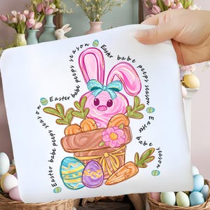 Może przedstawiać: Biała poszewka na poduszkę z motywem wielkanocnym. Przedstawia różowego królika z niebieską kokardą, marchewki i pisanki. Słowa "Easter babe peeps season" są napisane wokół wzoru. Poszewka jest trzymana przez dłoń.