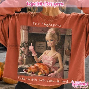May include: A rust-coloured crewneck sweatshirt featuring a graphic print of a blonde doll wearing a pink apron and gloves, holding a roasted turkey on a platter while giving the middle finger. The text reads It's Thanksgiving and Time to give everyone the bird.
