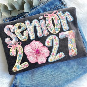 Puede incluir: Una sudadera gris oscuro con la inscripci&oacute;n "SENIOR 2021" en apliques florales y patchwork. El n&uacute;mero "2" est&aacute; emparejado con una gran flor rosa. Lazos rosas adornan las letras y los n&uacute;meros.
