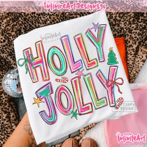 May include: A white t-shirt featuring the text HOLLY JOLLY in large, colorful, outlined letters. The design includes a green Christmas tree, a small candy cane, a yellow star, and two bows in green and red, all printed on the front of the shirt.